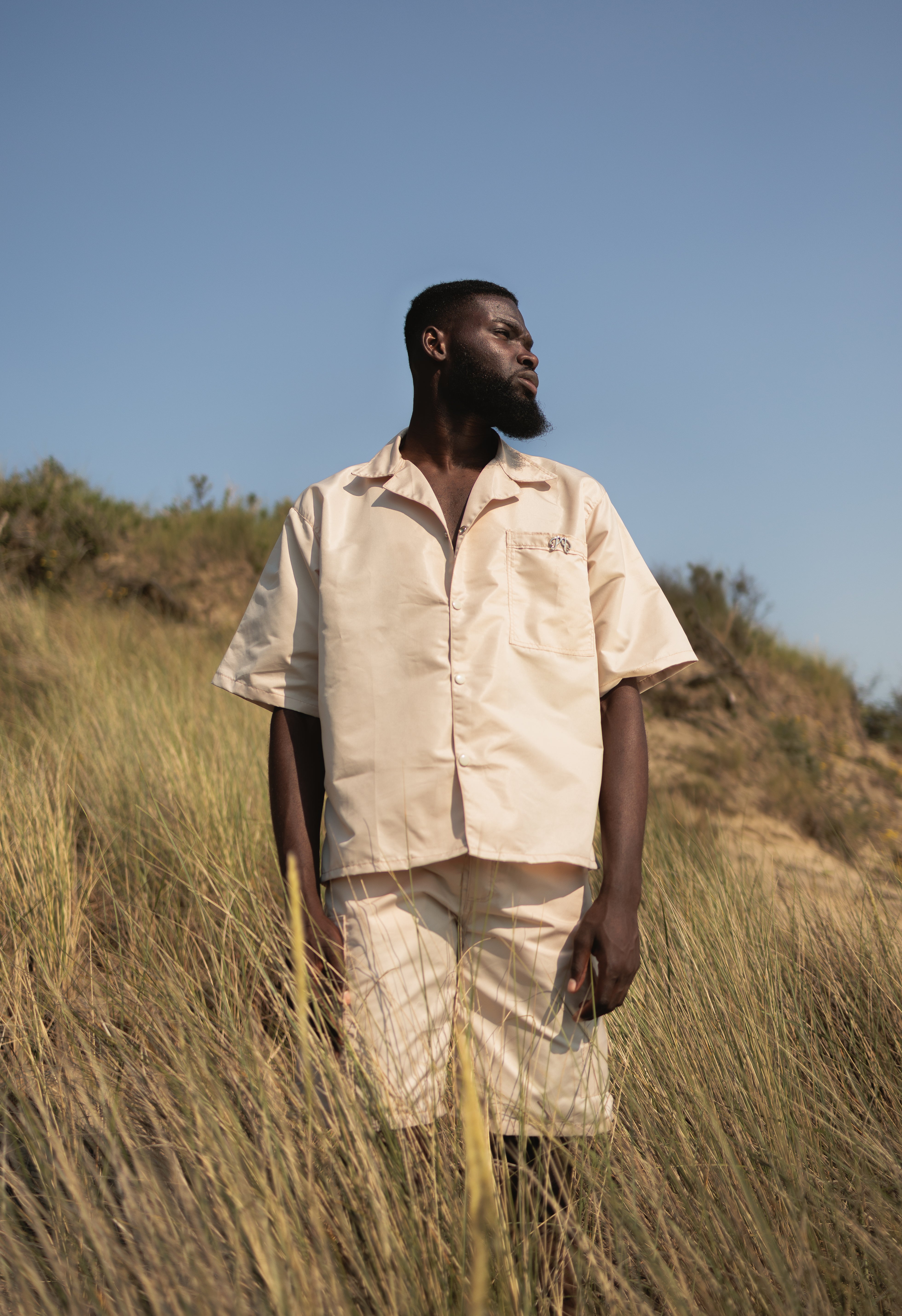 model wearing The Picou Nylon shirt in beige/cream is a short-sleeve button-up with a notched collar and a front pocket on the left chest. The pocket features a small metal tag design with intertwined letters in silver. This casual shirt offers a relaxed fit and is crafted from lightweight fabric, ideal for fashion-forward individuals seeking quality.