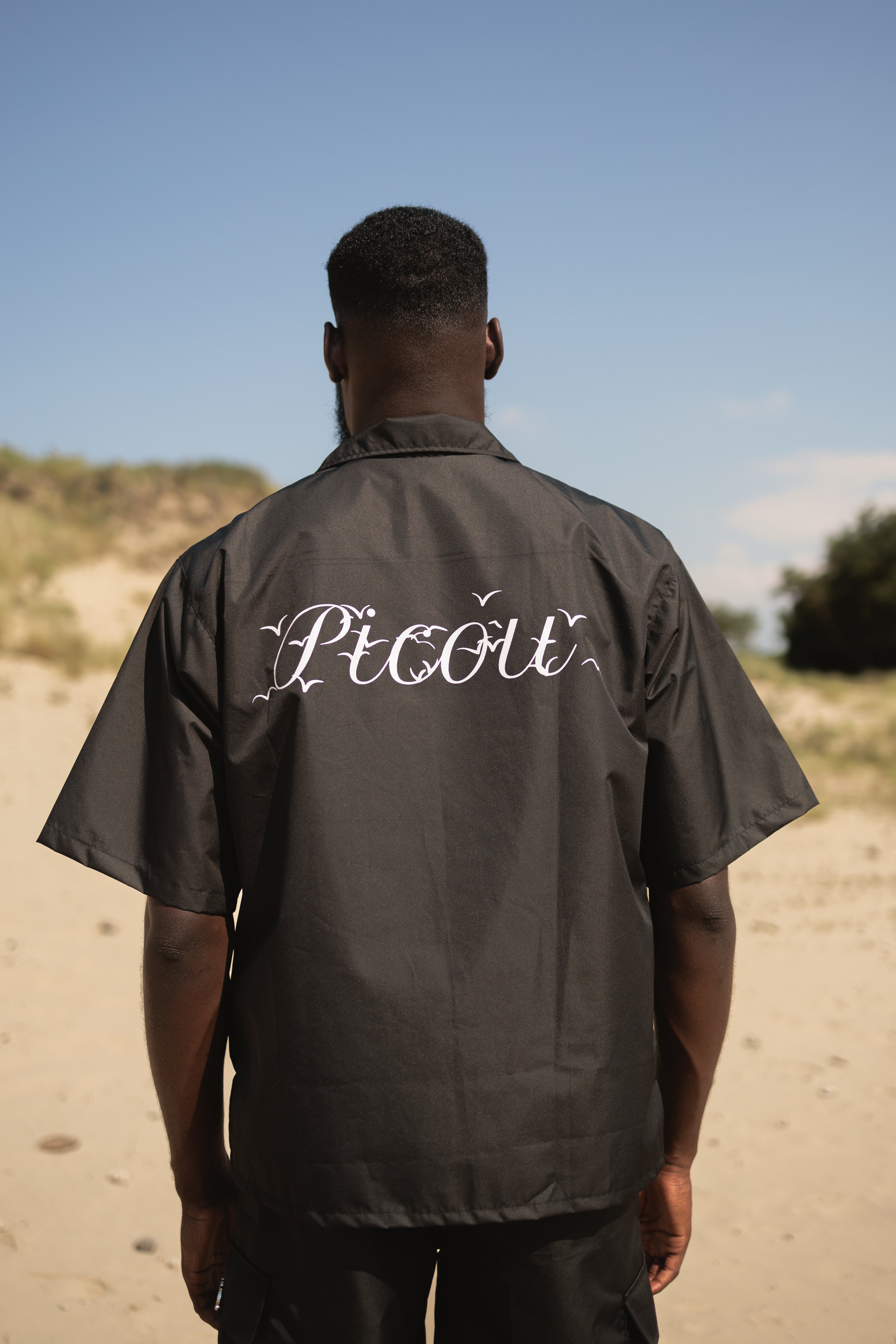 model wearing The Picou Nylon shirt in black is a short-sleeve button-up with a notched collar and a front pocket on the left chest. The pocket features a small metal tag design with intertwined letters in silver. This casual shirt offers a relaxed fit and is crafted from lightweight fabric, ideal for fashion-forward individuals seeking quality.