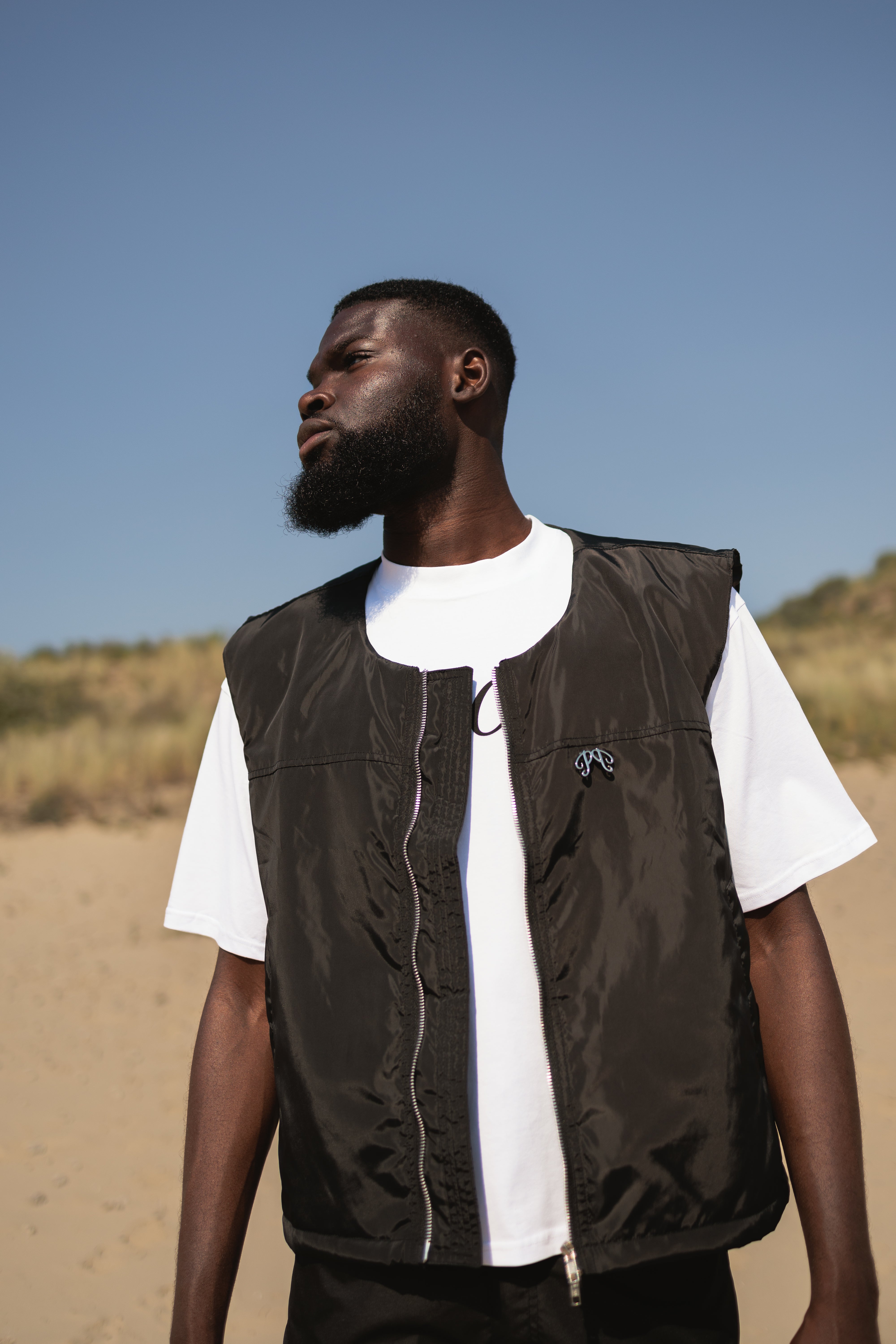 Picou Polyester bodywarmer in black exudes luxury and style with its shiny, lightweight fabric. This sleeveless zip-up vest features a high collar, two side pockets, and a small metal logo on the left side of the chest. The silver zipper adds a touch of contrast and quality to the overall black design.