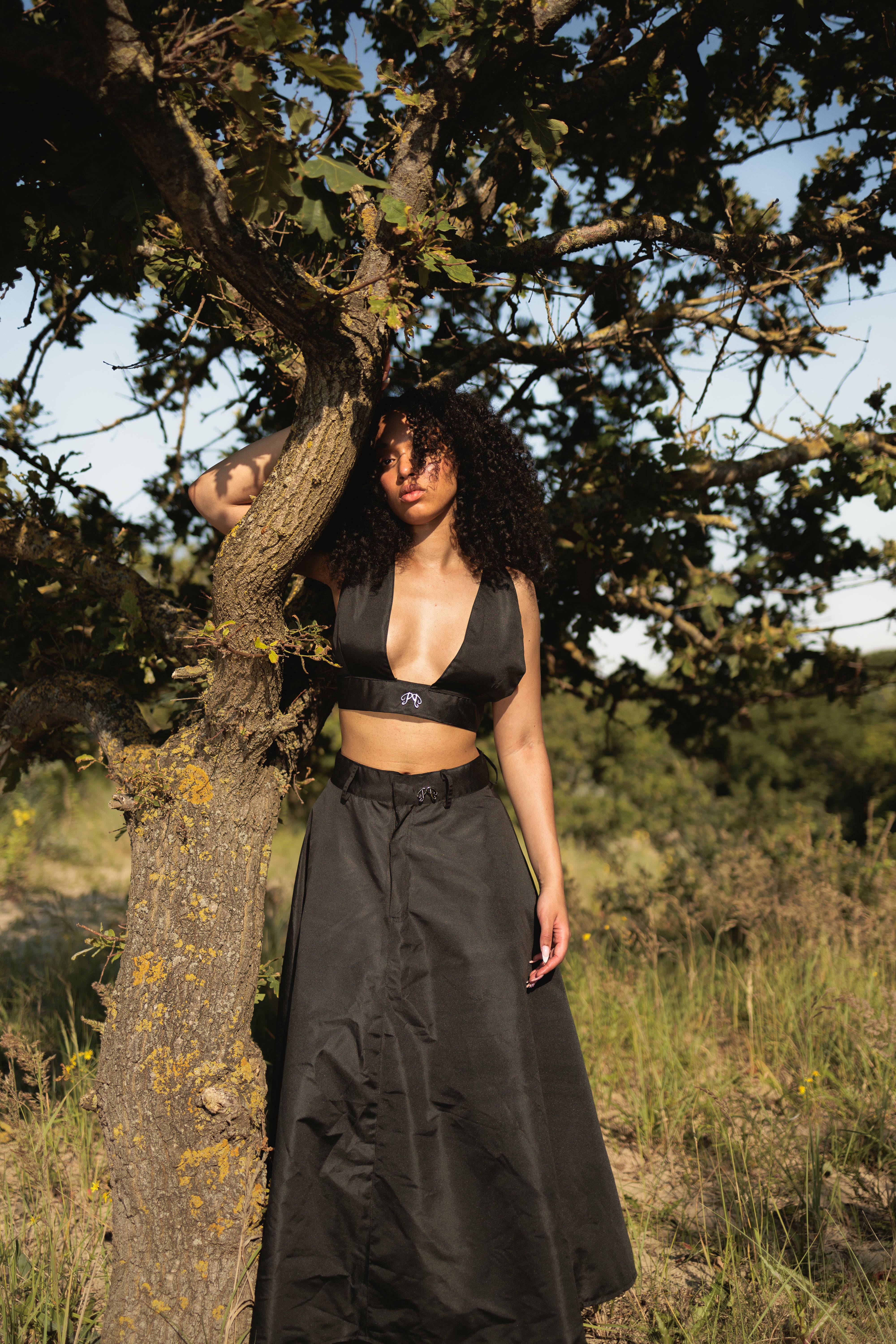 model wearing a Long, A-line black nylon skirt by Picou, featuring a high waist and a slight flare at the bottom. The smooth and slightly glossy fabric enhances its trendy style and quality, while a small buckle at the waist adds an extra touch of detail.