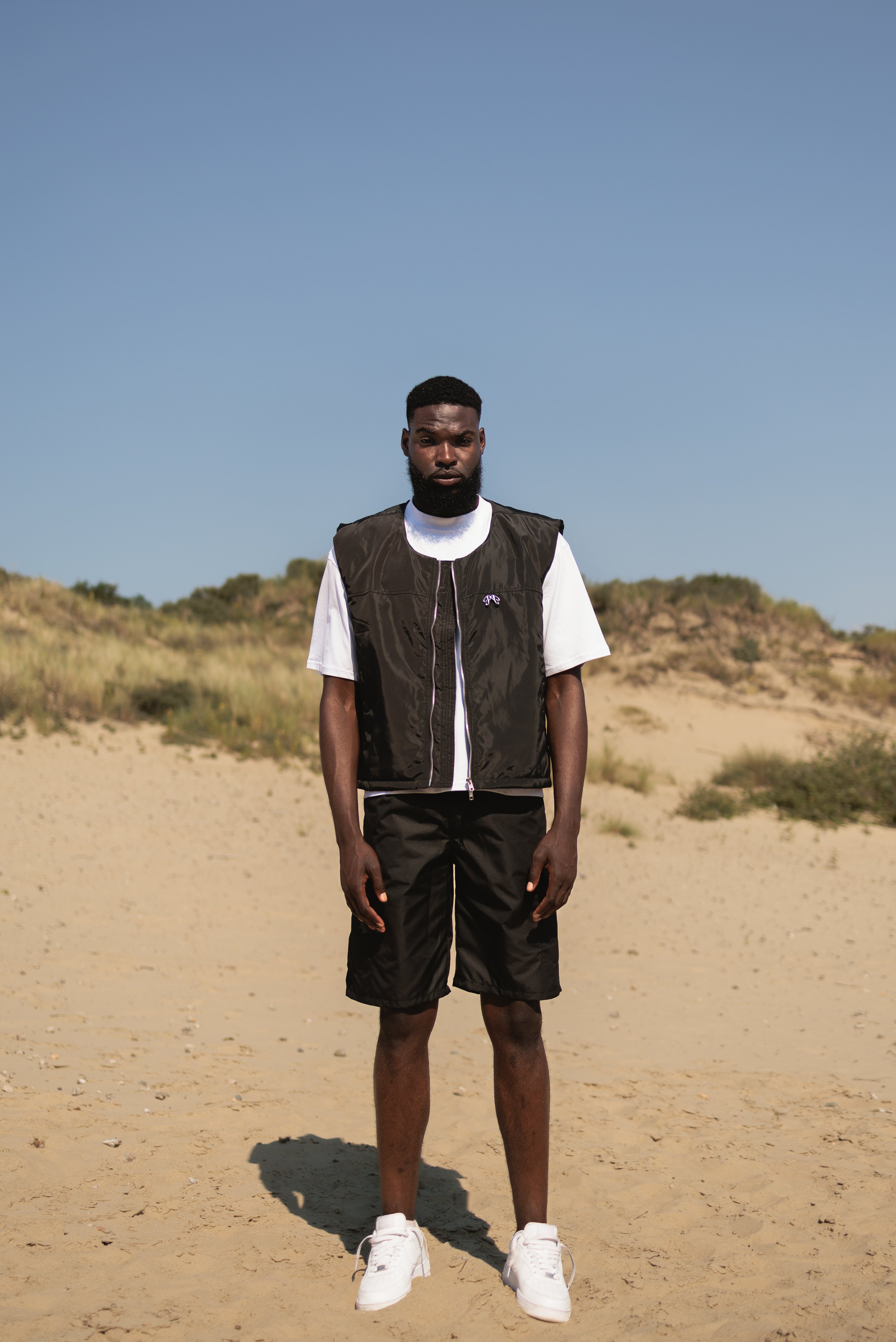 Picou Polyester bodywarmer in black exudes luxury and style with its shiny, lightweight fabric. This sleeveless zip-up vest features a high collar, two side pockets, and a small metal logo on the left side of the chest. The silver zipper adds a touch of contrast and quality to the overall black design.