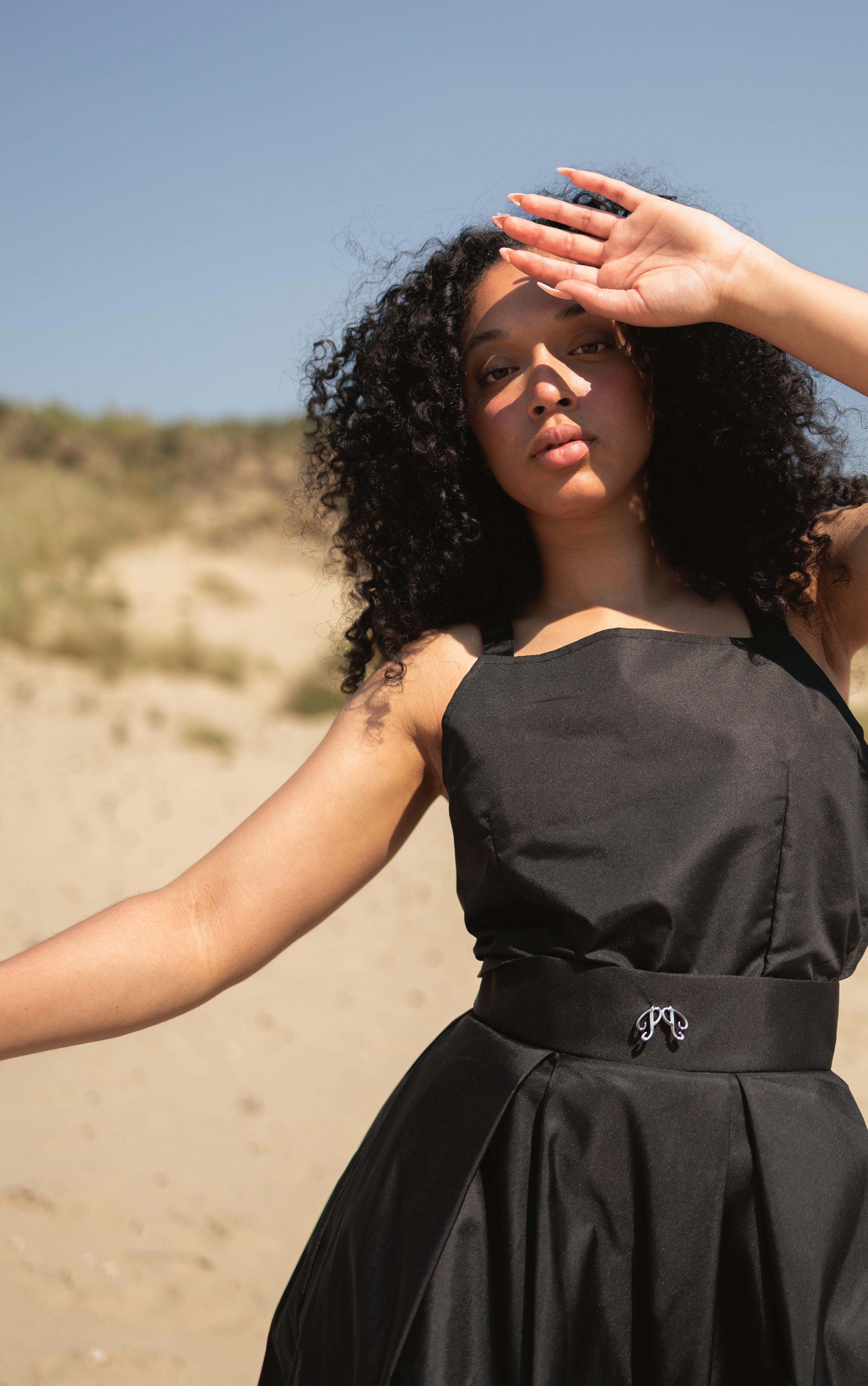 The Picou Nylon Dress is a black sleeveless piece featuring a square neckline and a wide waistband adorned with a subtle decorative clasp. It boasts a knee-length, pleated skirt that adds volume and texture. The smooth, slightly glossy fabric makes it perfect for achieving a casual yet chic style.