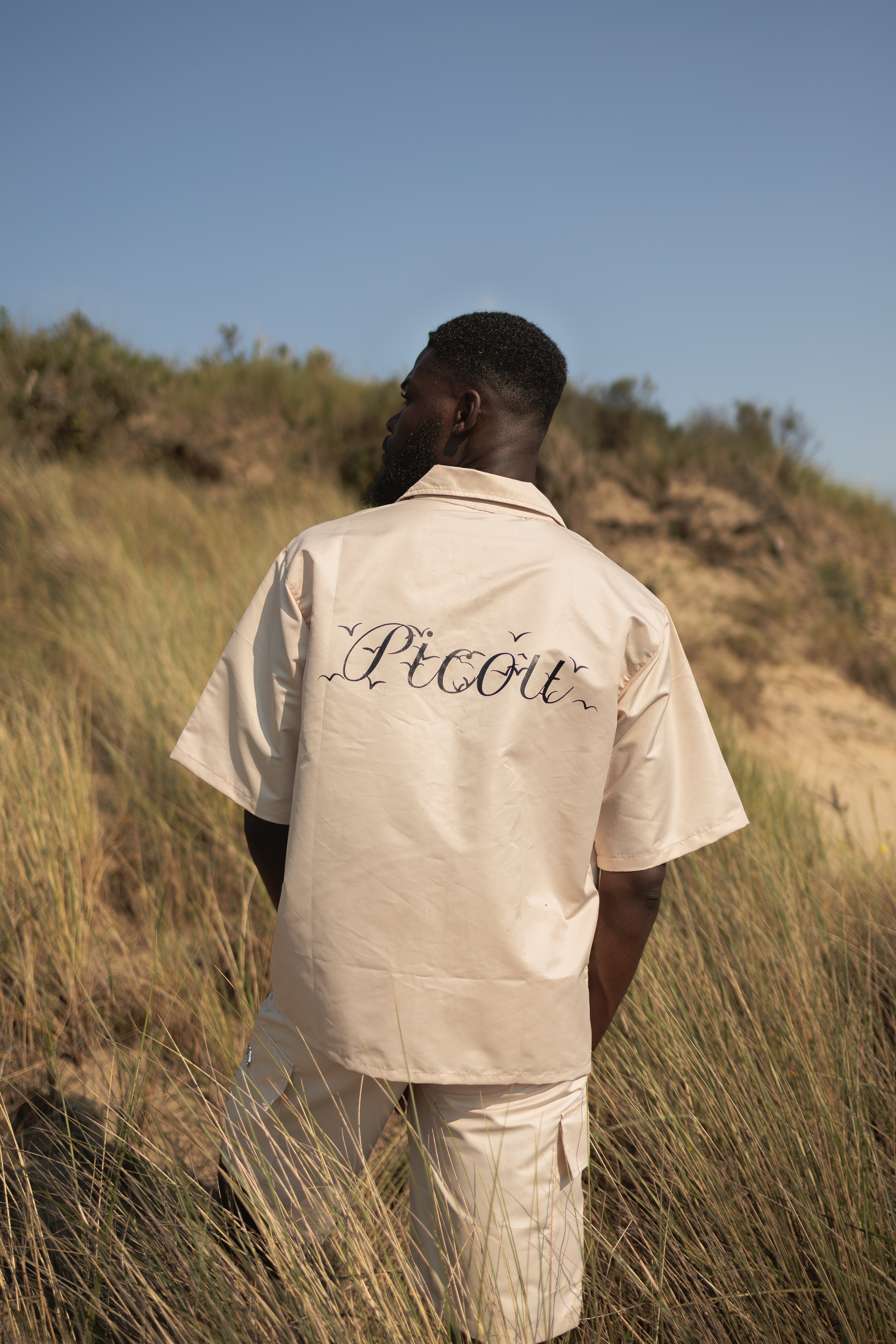 model wearing The Picou Nylon shirt in beige/cream is a short-sleeve button-up with a notched collar and a front pocket on the left chest. The pocket features a small metal tag design with intertwined letters in silver. This casual shirt offers a relaxed fit and is crafted from lightweight fabric, ideal for fashion-forward individuals seeking quality.